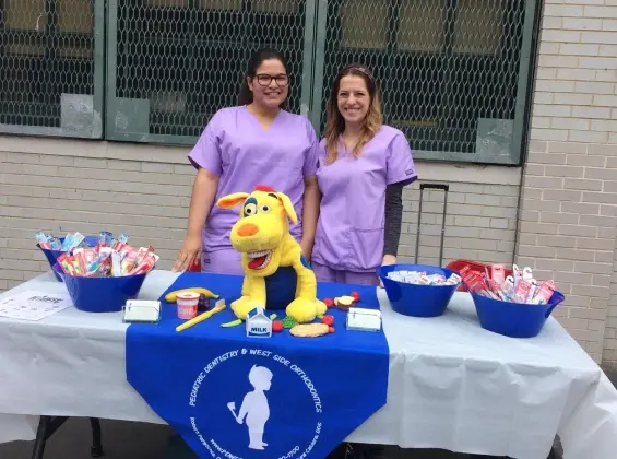 Two dental professionals in purple shirts at a charity event table with Big Bird mascot and supplies.